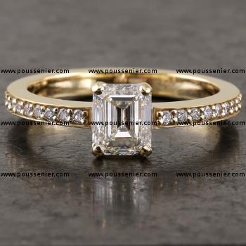 ring with a larger emerald cut diamond set with four prongs on a band with a rectangular profile pavé set with small brilliant cut diamonds