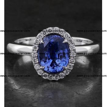 entourage ring with a central oval cut heated Ceylon sapphire surrounded by accent stones in castle pavé setting mounted on a bird-shaped or hollow V on a slightly convex band set with a small diamond on the side (compatible with a wedding ring)