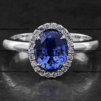 entourage ring with a central oval cut heated Ceylon sapphire surrounded by accent stones in castle pavé setting mounted on a bird-shaped or hollow V on a slightly convex band set with a small diamond on the side (compatible with a wedding ring)