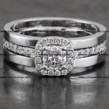  halo or entourage ring with a central cushion cut diamond surrounded with smaller brilliant cut diamonds on a castle-set band with palmettes between which a low mounted double V or pyramid shaped setting